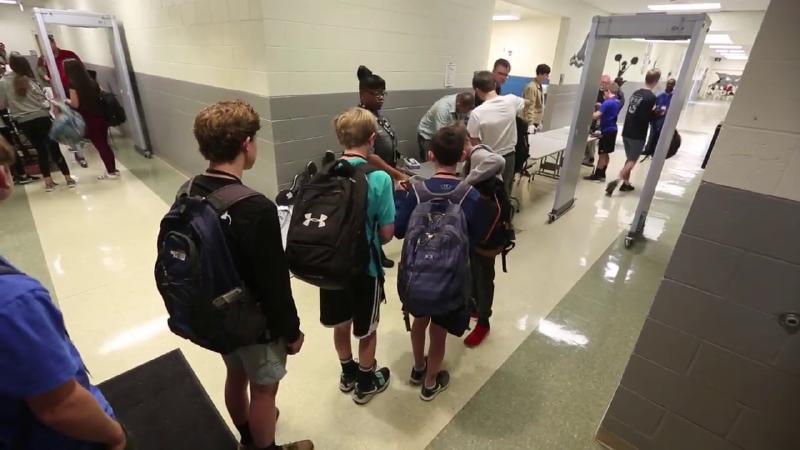 Young students between the ages of 10 and 12 stand in line at a metal detector, while wearing their backpacks.